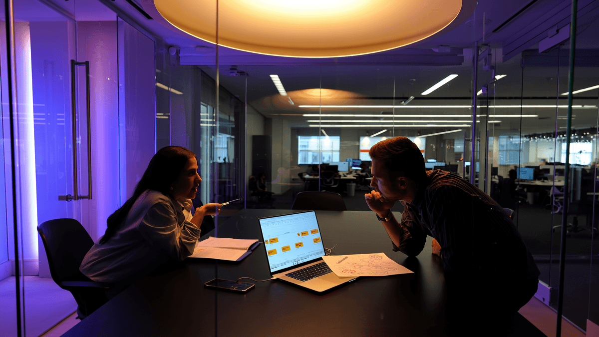 Two professionals in deep conversation through glass conference room wall with project timeline visible