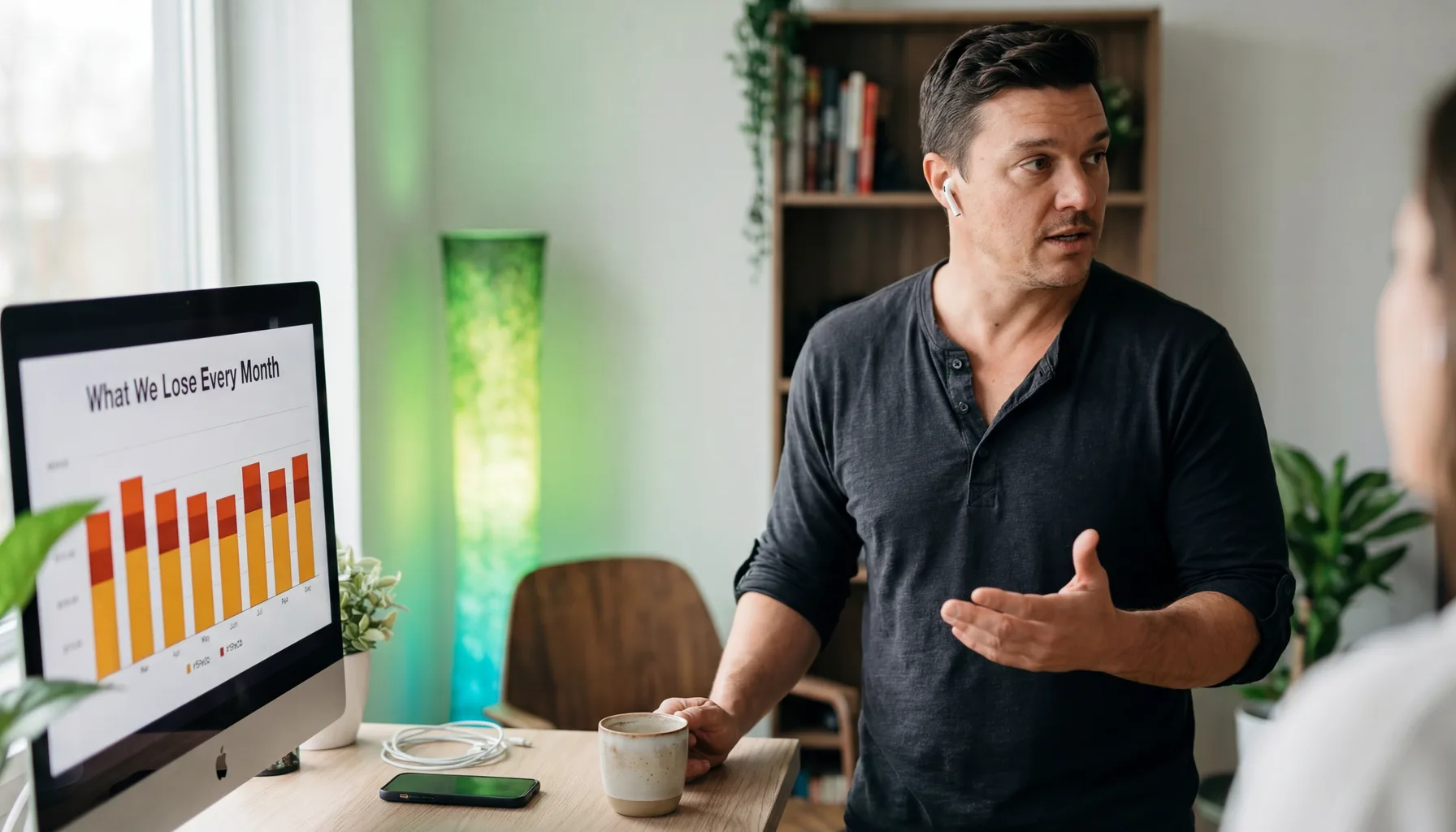 ADAM at standing desk gesturing mid-explanation, laptop showing cost-of-delay chart, amber and blue light